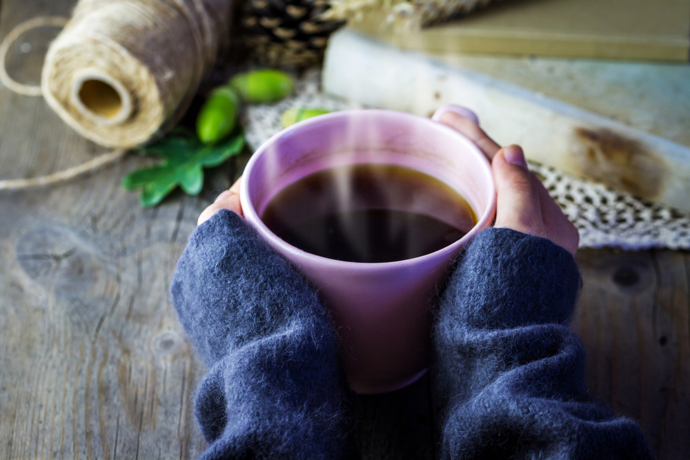 Close-up view of pink cup of hot coffee in female hand. Twine, old book and acorns on old wooden background