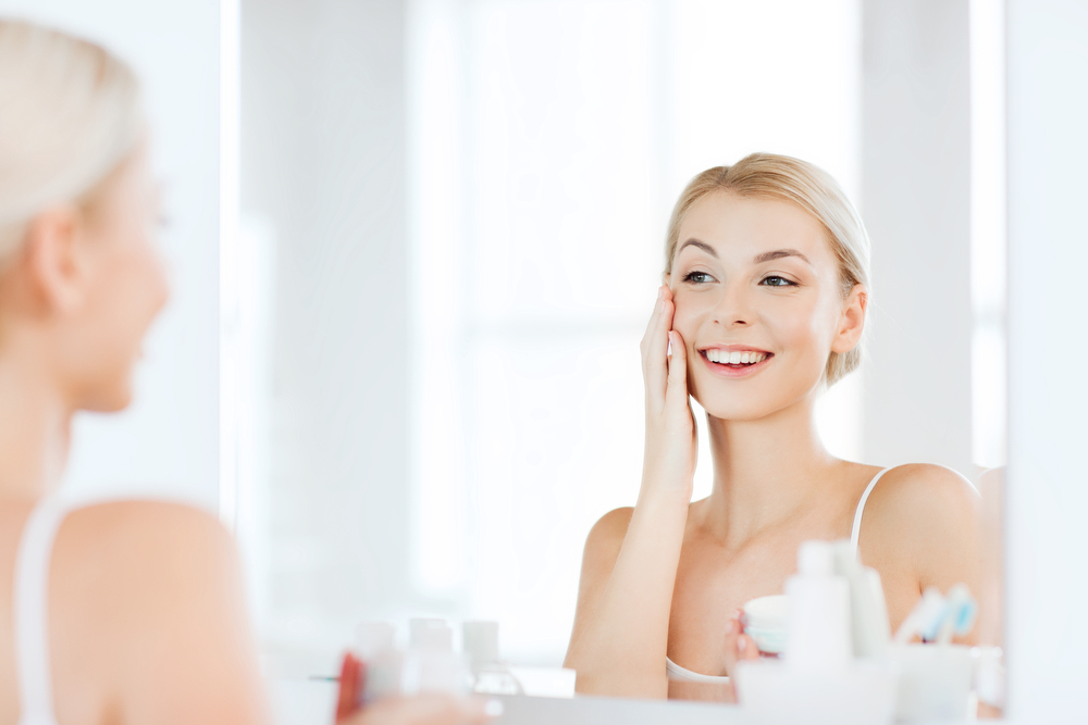 beauty, skin care and people concept - smiling young woman applying cream to face and looking to mirror at home bathroom