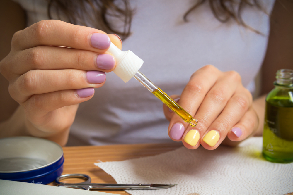 Female hand holding a pipette with oil. A young girl sits at a table and gets a yellow oil on the nails. On the table are nail clippers, a bottle of oil, a jar of cream and other nail accessories.