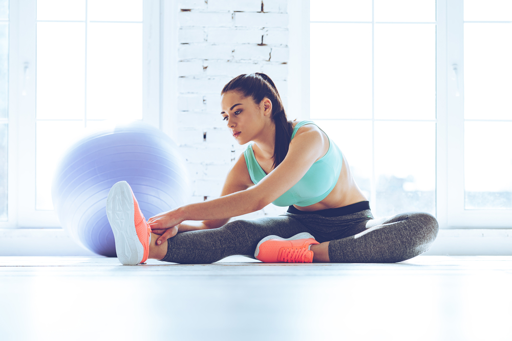 Great progress. Young beautiful woman in sportswear doing stretching while sitting on the floor in front of window at gym