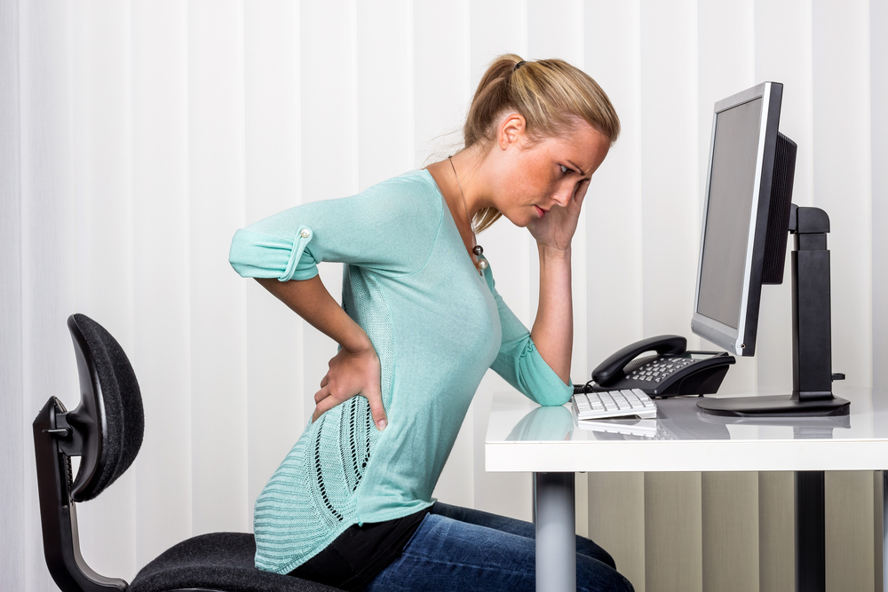 a woman sitting at a desk and has pain in the back. symbol photo for proper posture at work in the office.