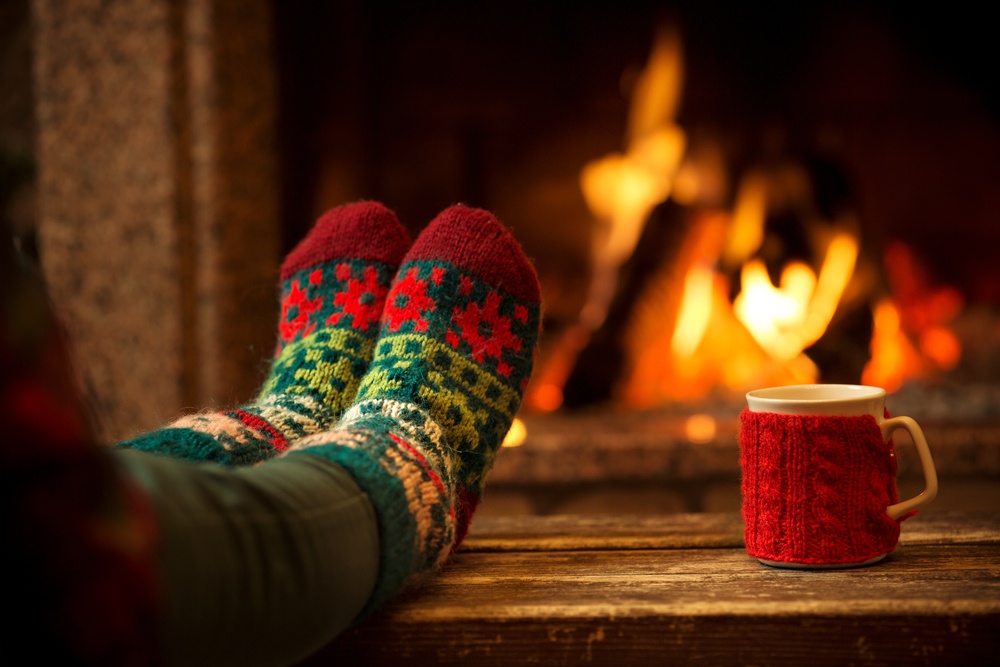 Feet in woollen socks by the Christmas fireplace. Woman relaxes by warm fire with a cup of hot drink and warming up her feet in woollen socks. Close up on feet. Winter and Christmas holidays concept.