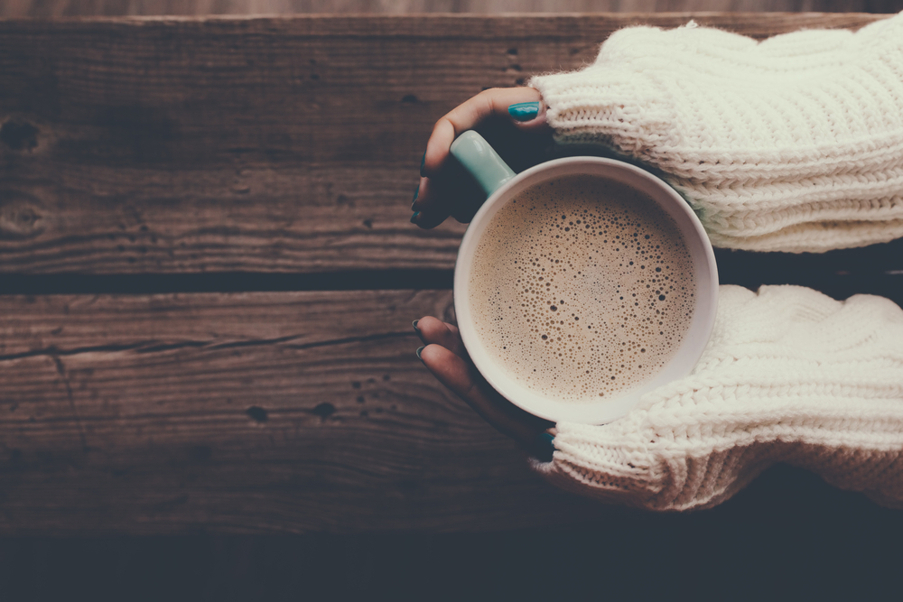 Woman holding cup of hot coffee on rustic wooden table, closeup photo of hands in warm sweater with mug, winter morning concept, top view